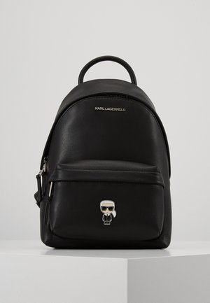 Mini-sac à dos en cuir noir avec poche zippée à l’avant, logo Karl Lagerfeld et petite épingle personnage portant des lunettes de soleil sur une surface blanche.