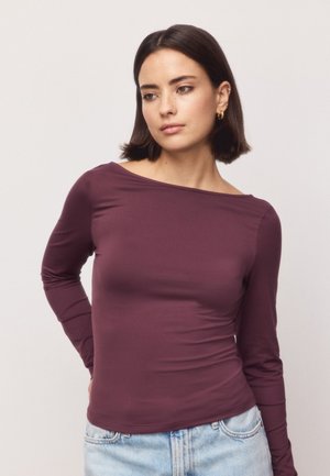 Femme aux cheveux foncés mi-longs, portant des créoles dorées et un haut ajusté à manches longues bordeaux, debout avec une main derrière le dos.