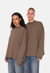 Langärmlige braune Baumwoll-T-Shirts mit lockerer Passform, die ein kleines gesticktes Logo auf der Brust und gerippte Abschlüsse aufweisen. Getragen von zwei Personen.