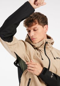 Vedenkestävä takki beige ja musta, jossa on huppu. Ominaisuudet vetoketjut ja säädettävät hihansuut. Sisäkerros näkyvissä, ja se esittelee tummanvihreän rakenteen.