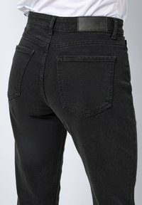 Jean en denim noir avec taille haute, cinq poches et texture lisse. Présente une patch de la marque à la taille et une coupe fuselée.
