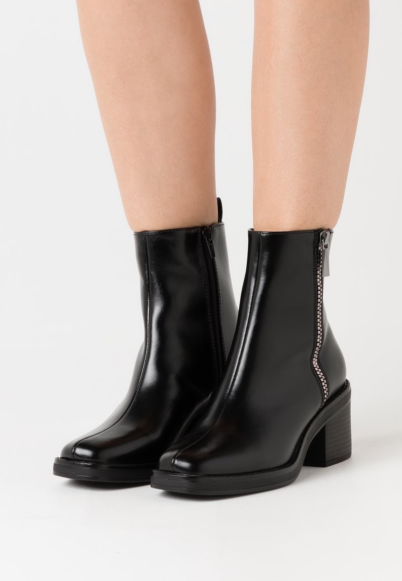 Bottes noires en cuir brillant à cheville avec fermetures éclair latérales et talons blocs, portées sur des jambes nues sur fond blanc.