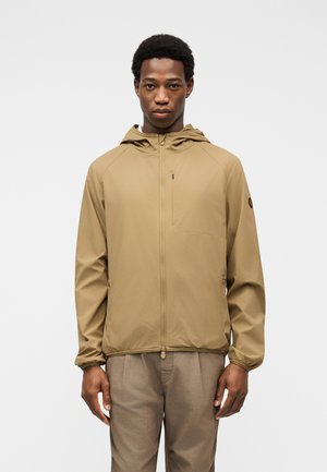 Mand iført en beige hættetrøje med lynlås og matchende bukser, stående mod en ensartet lysegrå baggrund, vendt mod fronten.