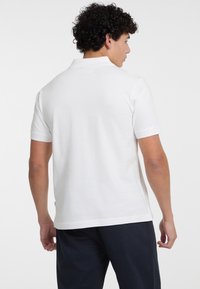 Camisa polo blanca de manga corta, con tejido suave, corte ajustado y cuello acanalado. Detalle de costura visible en el dobladillo y en los puños.