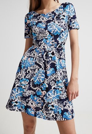 Vrouw draagt een knielange, kortemouwen jurk met een blauw, wit en lichtroze bloemenpatroon op een donkere achtergrond.
