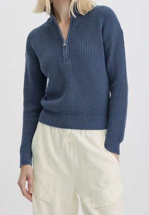 Femme portant un pull en tricot bleu à demi-fermeture éclair et un pantalon cargo crème, une main dans la poche, sur un fond clair uni.