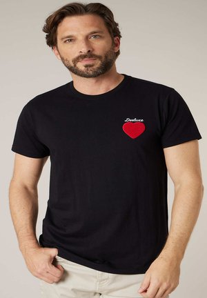 Homme barbu portant un t-shirt noir avec un patch cœur rouge et le texte « Deeluxe » sur la poitrine, debout devant un fond uni.