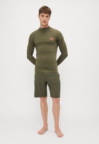 Ilga rankovė rash guard ir šortai, abu olivinės spalvos. Rash guard turi mažą logotipą ant krūtinės ir puoštą prigludusiu dizainu. Lygi tekstūra.