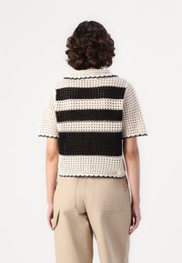 Gestreiftes, kurzärmliges Strickoberteil in Creme und Schwarz, mit durchsichtiger Netzstruktur, horizontalen Streifen, Wellenkragen und Saum. Kombiniert mit beigen Hosen.
