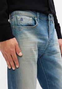 Ανοιχτό μπλε denim τζιν με σταδιακό εφέ ξεθωριάσματος, διαθέτει μπροστινή τσέπη με μπλε εμβλημα και αντίθεση ραφής κατά μήκος των ραφών.