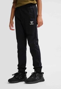 Schwarze Sporthosen mit einem Kordelzug-Bund, seitlichen Taschen und einem weißen Logo am Oberschenkel. Kombiniert mit schwarzen Turnschuhen.