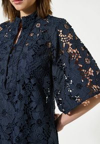 Vestido de encaje azul marino con diseños florales, cuello alto, cierre de botones en la parte delantera y mangas hasta el codo con detalles recortados.