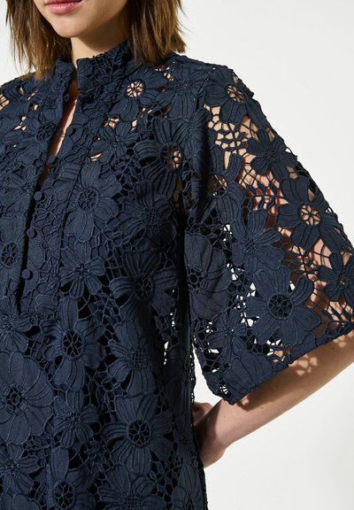 Femme portant un haut en dentelle florale bleu marine avec manches courtes et col mandarin, affichant des motifs de fleurs découpés complexes.