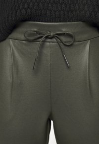 Pantalons couleur vert foncé, taille haute, avec un lien à l'avant et des plis. Fabriqués en tissu lisse, avec un design épuré et moderne sans poches visibles.