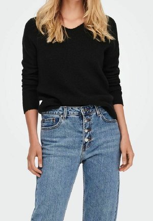 Vrouw draagt een zwarte trui met lange mouwen, ingestopt in een high-waist blauwe spijkerbroek met zichtbare knopen aan de voorkant.