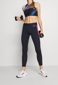 Svart sport-bra med blå detaljer, åtsittande marinblå leggings med rosa sidosträck, och vita gympaskor med randiga detaljer.