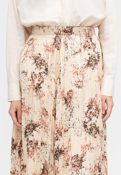 Person wearing a pleated skirt with pink and black floral print and a tucked-in long-sleeve cream blouse, hands resting at sides.