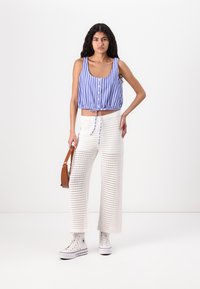 Blau-weißer gestreifter, taillierter Tanktop mit Knöpfen, weiße weit geschnittene Hosen aus offenem Strick und weiße High-Top-Sneaker. Hält eine braune Tasche.