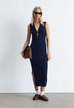 Marineblaues, geripptes ärmelloses Kleid mit tiefem V-Ausschnitt und Seitenschlitzen, goldene Hardware-Akzente am Ausschnitt, kombiniert mit einer braunen Tasche und braunen Schuhen.