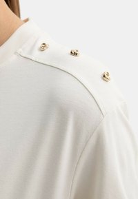 Épaule d'une chemise blanche avec trois petits boutons dorés en forme de nœud le long de la couture près du col.