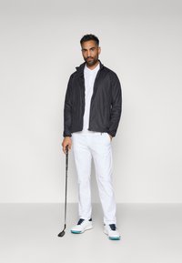 Melns viegls jakas ar rāvējslēdzēju, balts polo krekls, pieguļošas baltas bikses un baltas golfa kurpes ar zilām akcentiem; tur rokā golfa nūju.