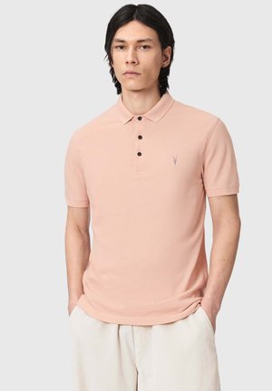 Junger Mann mit dunklen Haaren, der ein hellrosa kurzärmeliges Poloshirt und hellbeige Hosen trägt, die Hände in den Taschen, neutraler Hintergrund.