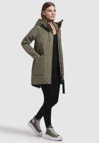 Parka vert olive avec capuche, fermeture éclair à l'avant et boutons-pression. Doté de deux grandes poches latérales et d'un dos légèrement plus long. Associé à des baskets montantes vertes.