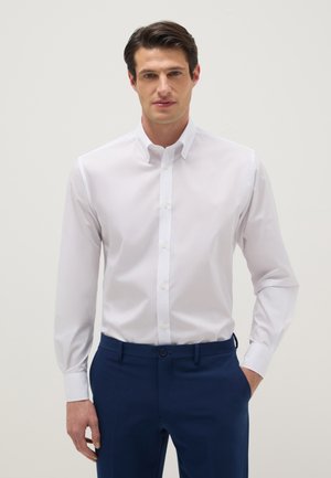 Mann trägt ein ordentlich zugeknöpftes weißes Hemd und dunkelblaue Hose, steht mit einer Hand in der Tasche vor einem schlichten Hintergrund.