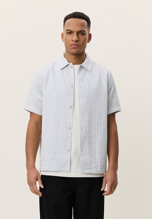 Homme portant une chemise boutonnée à manches courtes texturée gris clair par-dessus un t-shirt blanc et un pantalon noir, debout devant un fond beige uni.