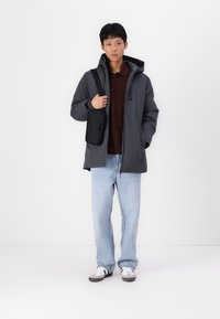 Mørkegrå dunjakke med hætte, lynlås foran og sidelommer. Båret over en brun sweater, parret med lyseblå jeans og hvide sneakers.