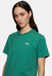 Groene katoenen t-shirt met een ronde hals en korte mouwen. Heeft een klein wit en rood "FILA" logo op de borst. Soepele textuur.