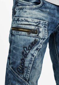 Jeans in denim con una tonalità blu sbiadita. Le caratteristiche includono una tasca con zip e cuciture decorative. Tessuto strutturato con un aspetto usurato.