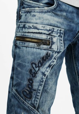 Jean en denim avec un délavage bleu. Comprend une poche zippée et des coutures décoratives. Tissu texturé avec une apparence usée.