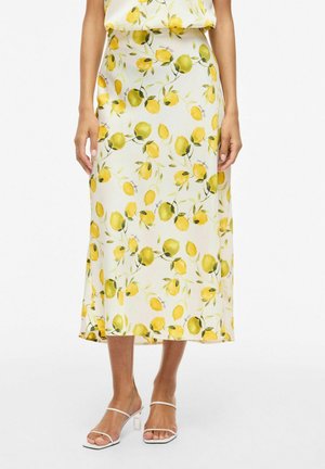 Biela midi sukňa so žltým vzorom citrónov a zelených listov, nosená s bielymi sandálmi na opätku, zobrazená od pása po nohy.