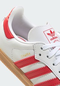 Bijela kožna tenisica s crvenim prugama, teksturirana guma potplata, s vezom "SAMBA" u zlatnoj boji, klasični adidas logo na jeziku.