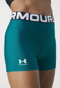 Shorts de compression Teal Under Armour avec une ceinture noire et blanche brandée à la taille et sur le haut des cuisses d’une personne.