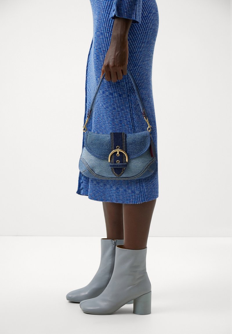 Personne à la peau foncée en robe bleue côtelée tenant un petit sac à main en denim avec une boucle dorée, portant des bottines gris clair à talons blocs.