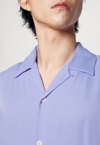 Camicia collare lilla realizzata in tessuto liscio con chiusura a bottone, caratterizzata da una leggera lucentezza e un design classico.