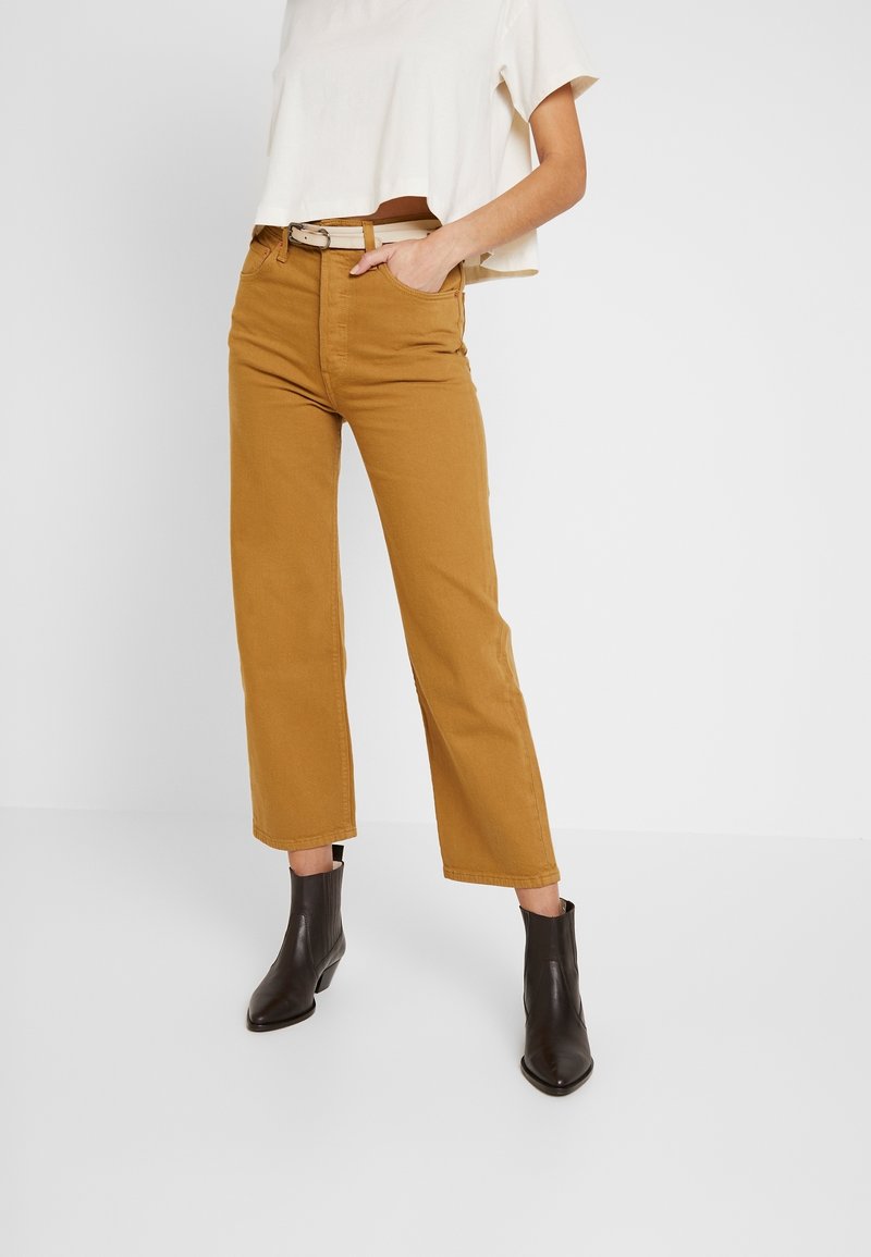 Personne portant un pantalon droit jaune moutarde, une ceinture beige, un top court blanc à manches courtes et des bottines noires, debout sur un sol blanc.