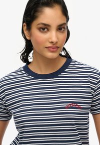 Camiseta de rayas azul marino y blanca con cuello redondo. Presenta un logo bordado en rojo de "Superdry" en el lado izquierdo del pecho. Material de algodón, mangas cortas.