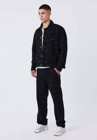 Giacca di jeans nera con motivo sottile, bottoni frontali e tasche, abbinata a pantaloni neri coordinati. Calzature sono sneakers grigio chiaro.