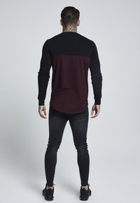 Man med tatuerad nacke och händer som bär en svart och mörkröd långärmad skjorta, svarta skinny jeans och svarta sneakers, vänd bort.