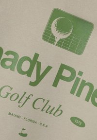 Tkanina kremaste boje s zelenim natpisom: "Shady Pines Golf Club." Uključuje grafiku golf loptice i tee-a, s detaljima "1979" i "Miami, Florida, SAD."