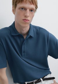Námořnická modrá polo košile s žebrovanou texturou, s límečkem a zapínáním na tři knoflíky, v kombinaci s černým pleteným pásem a bílými kalhotami.