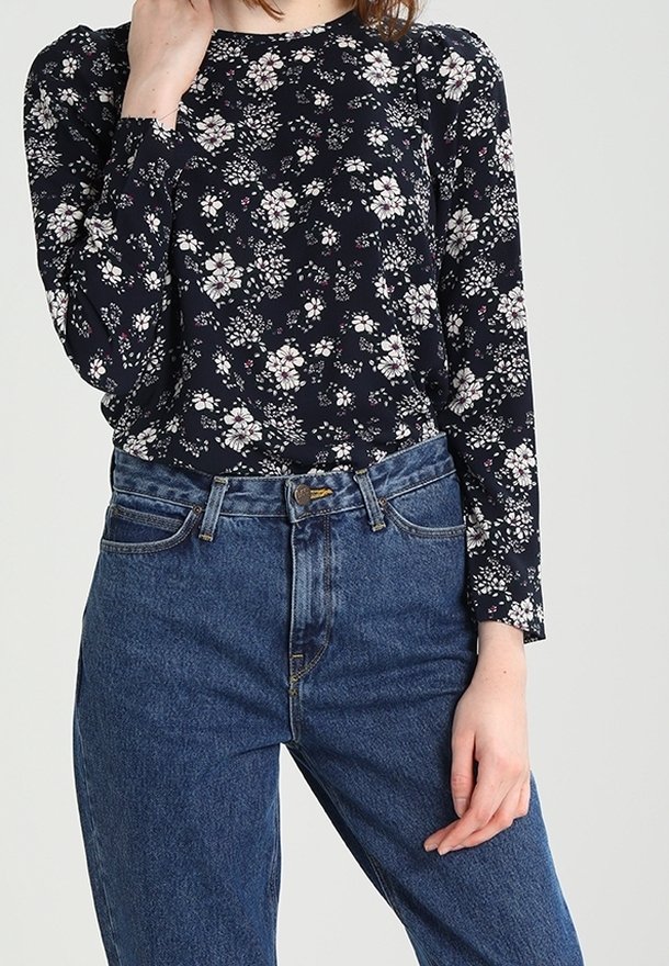 Mörkblå långärmad topp med vitt blommönster, rund halsringning och elastiska muddar, kombinerad med högmidjade blå jeans med knäppning framtill.