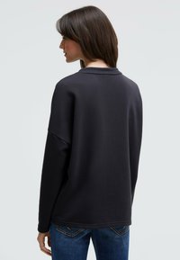 Schwarzer Sweatshirt mit lockerer Passform, rundem Ausschnitt und überschnittenen Schultern. Weicher Stoff und gerippte Bündchen, kombiniert mit blauen Jeans.