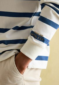 Pasiasta koszula z długim rękawem w niebiesko-białe paski, z prążkowanymi mankietami. Wyszywane "JOULES MCMLXXX" w złotym kolorze na mankiecie. Spodnie z sztruksu w kolorze beżowym.