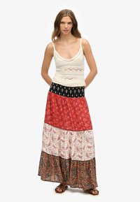 Gehaakte witte tanktop met verstelbare bandjes, gecombineerd met een lange, geprinte maxi-rok met rode, paisley- en bloemenmotieven.
