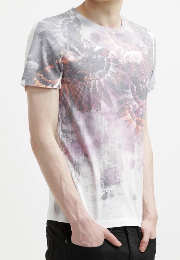 Homme portant un t-shirt blanc et gris avec un grand motif d'ailes d'oiseau, mains dans les poches, et pantalon noir.