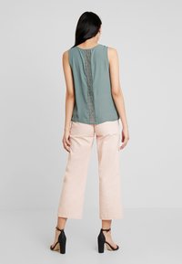 Top senza maniche verde smorzato con dettagli in pizzo al centro, abbinato a pantaloni ampi rosa chiaro e sandali neri con tacco.